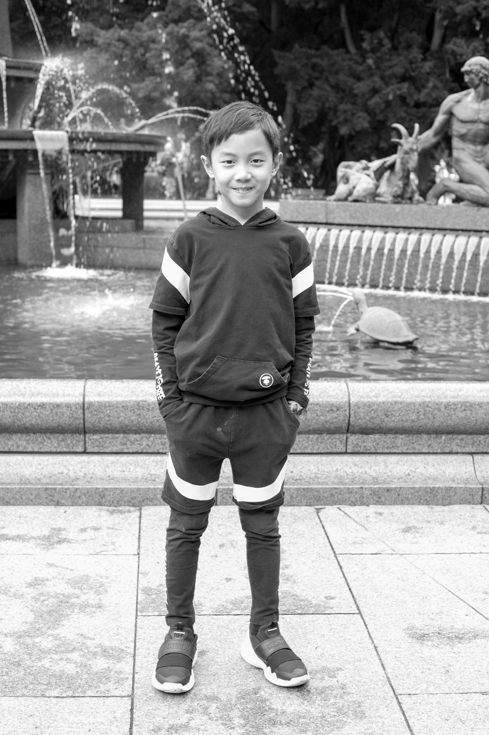 Black and white photo of young boy in sports gear in front of fountain.