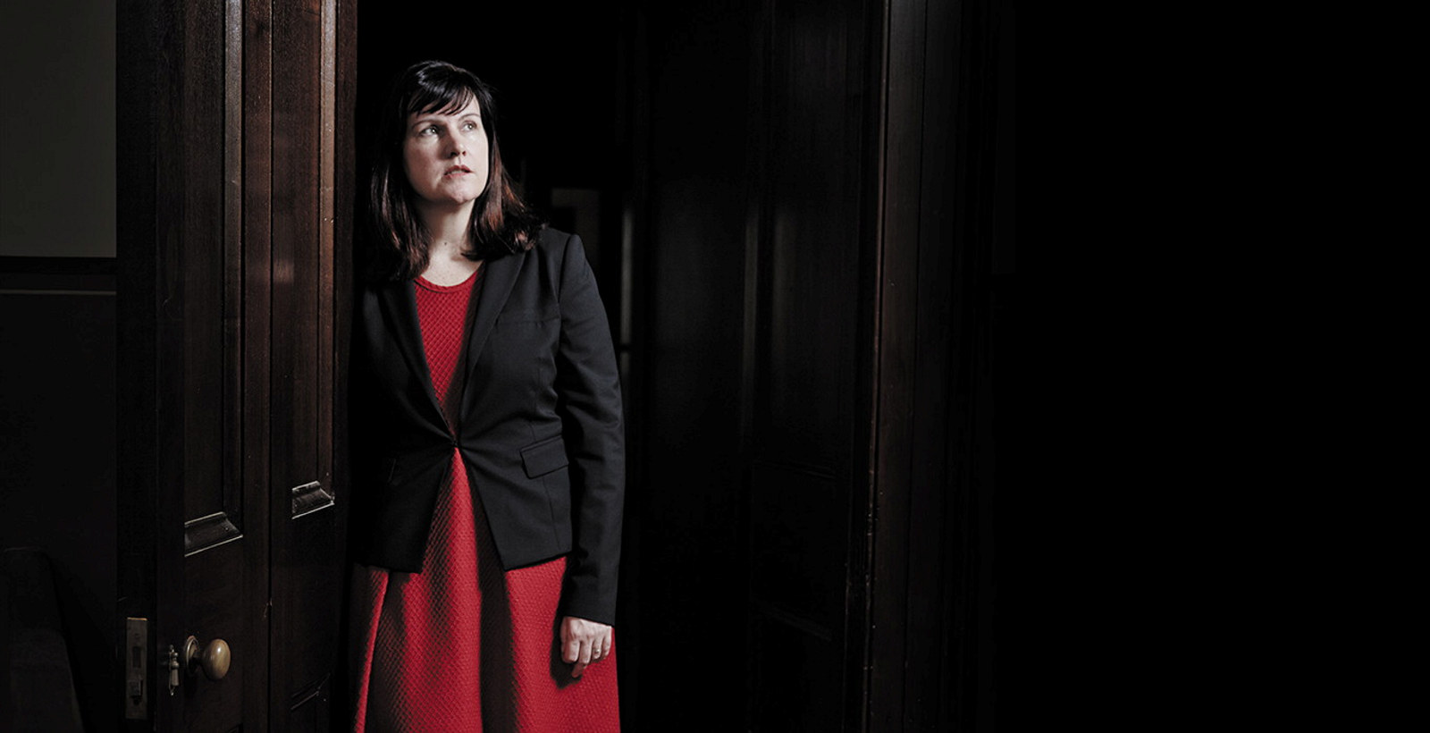 Dark haired woman wearing a red dress and black blazer leaning on a wooden door frame