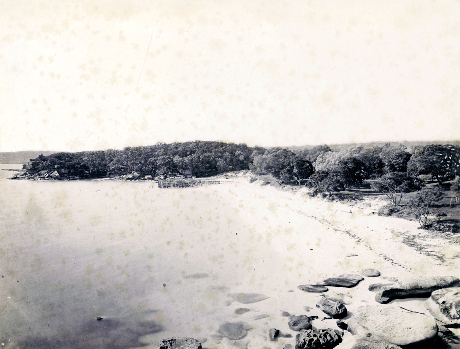 B/W photo showing a beach-lined bay with rocks and some kind of makeshift swimming enclosure at the water's edge.