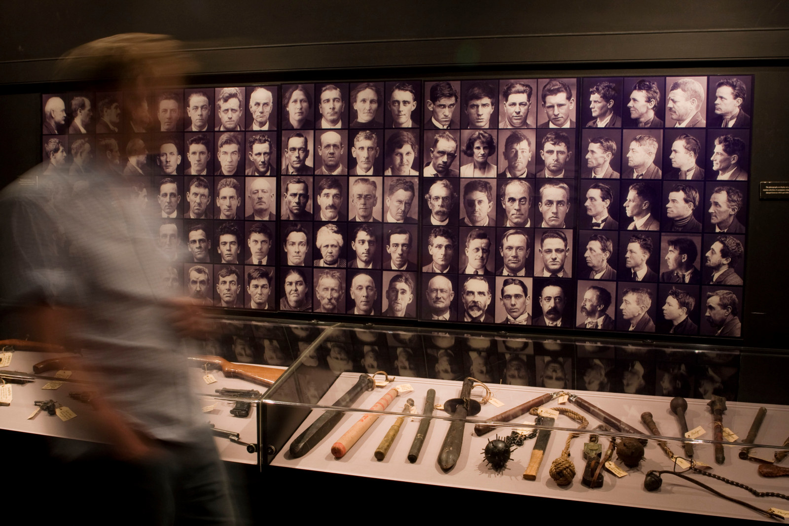 Photo showing person rushing past a photographic display of portraits and weapons under glass in a showcase.