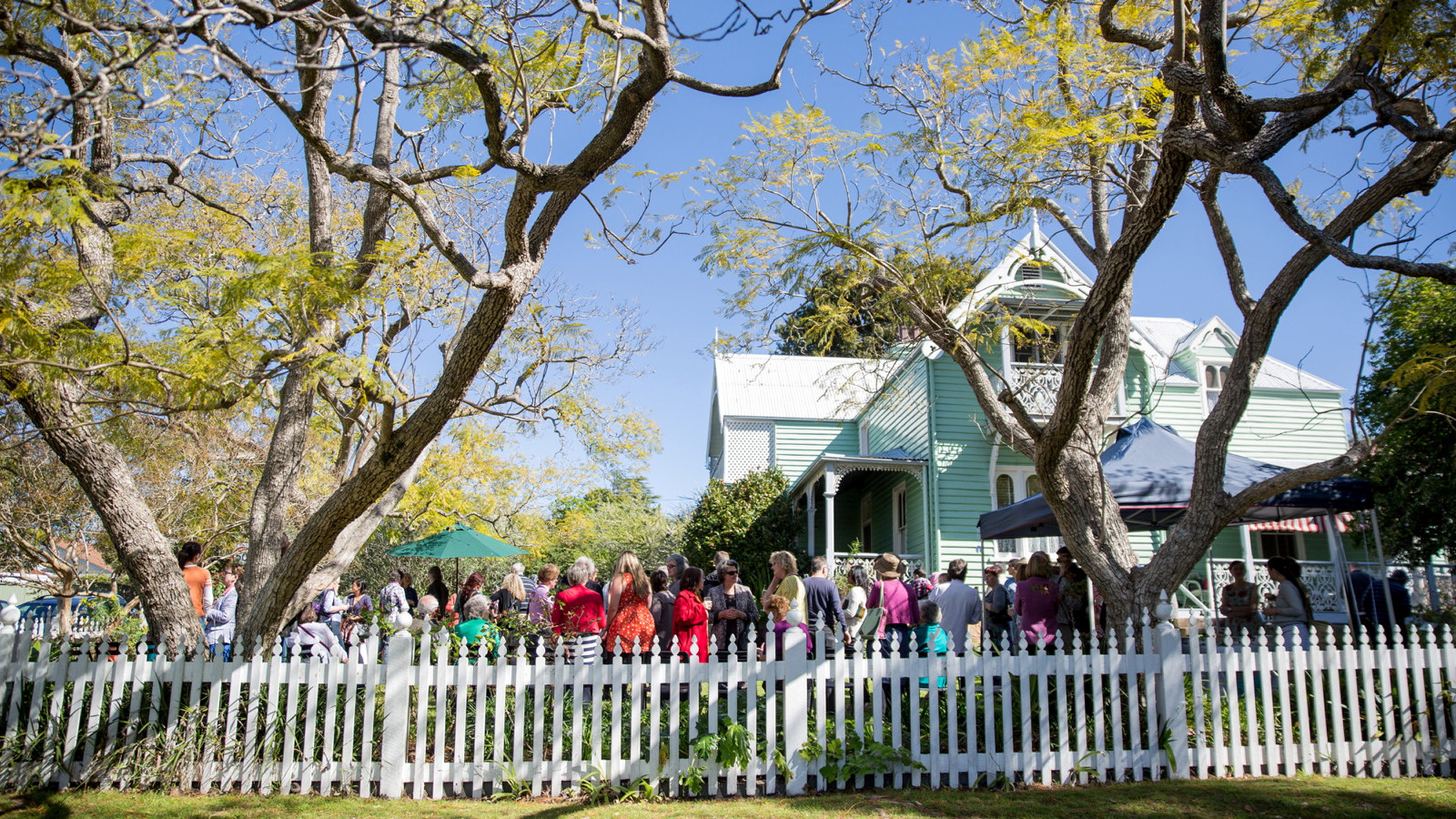 Large gathering in garden around wooden house, surrounded by white picket fence.