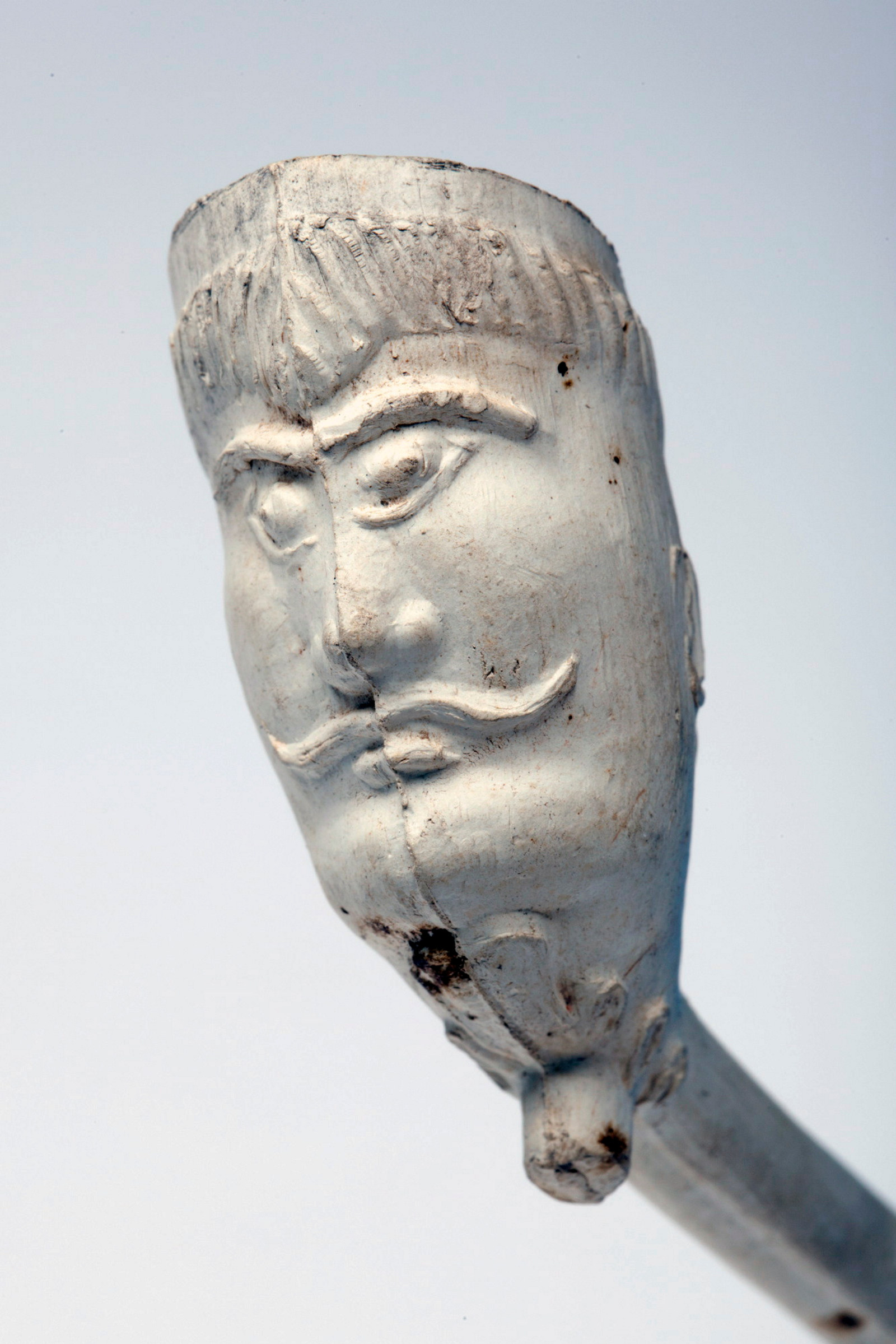 Front view of face carved onto white clay pipe bowl. Face has prominent eyebrows and moustache.