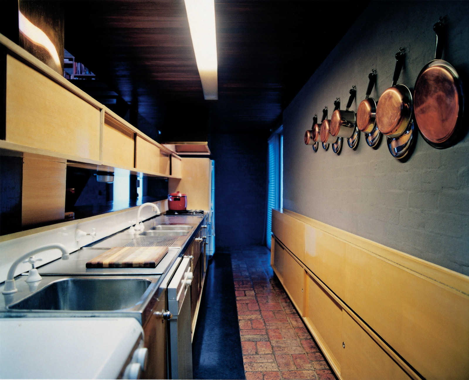 This is a colour photograph of a galley-style kitchen with dark blue walls, timber panelled cupboards, and kitchen pots hanging along one wall