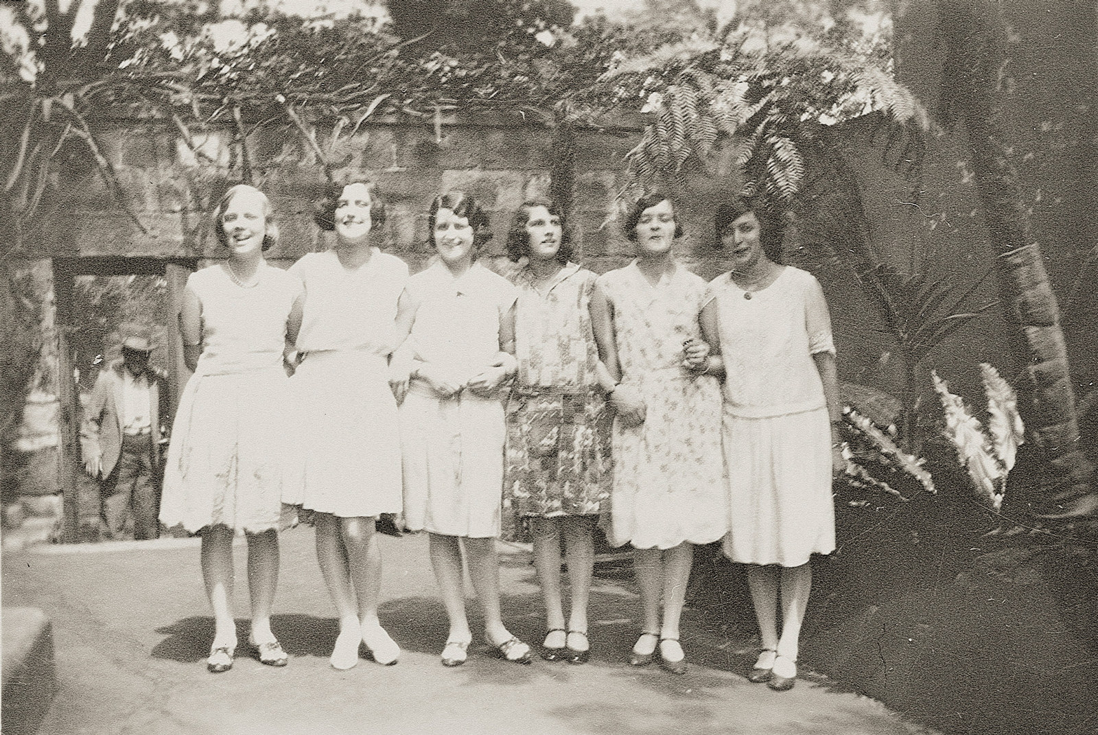 A group portrait of 6 women standing in front of a stone wall with shrubs growing over it. The women have linked arms and are all smiling at the camera.