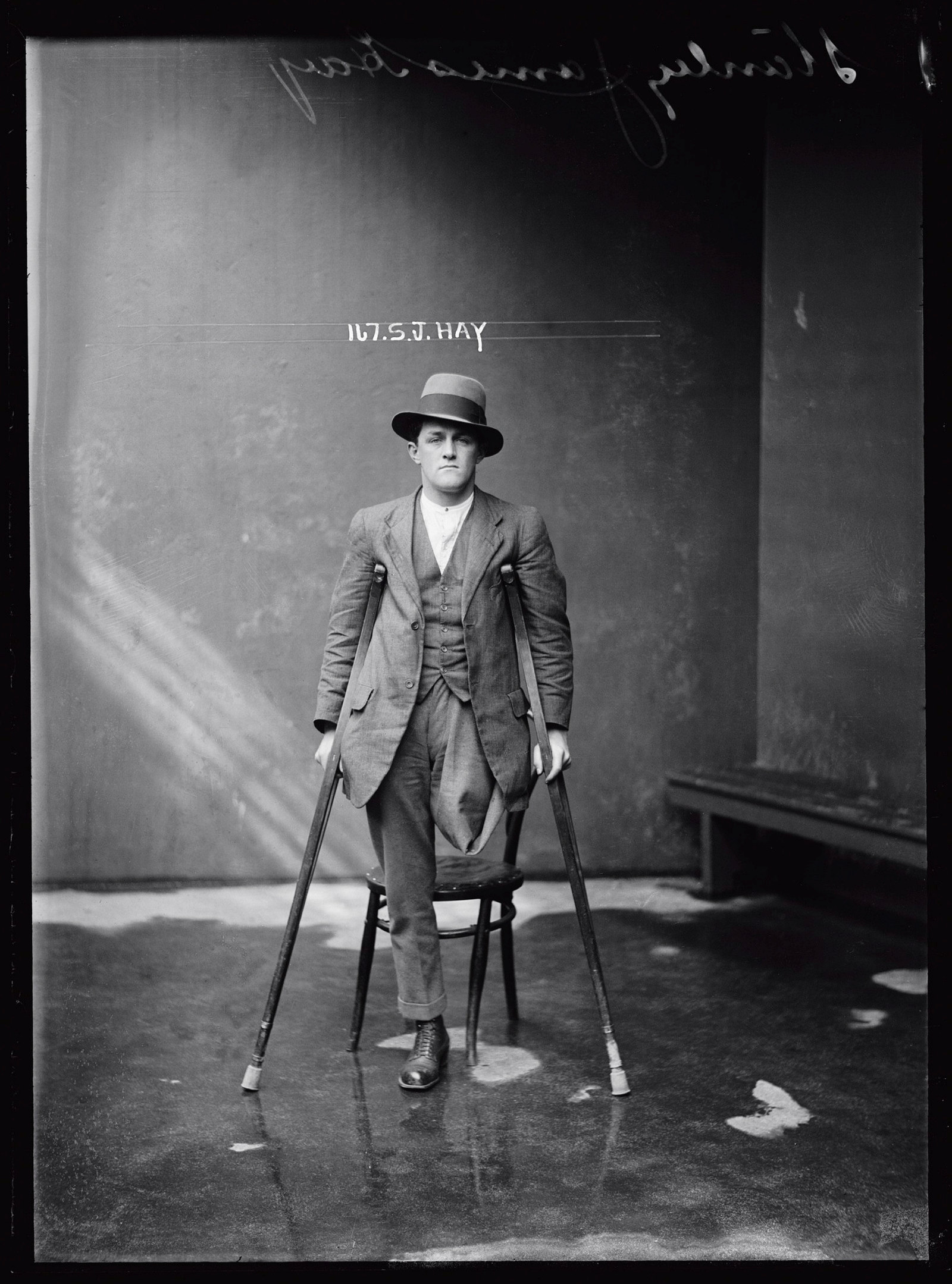 Black and white mugshot of one-legged man standing with crutches and wearing a hat.