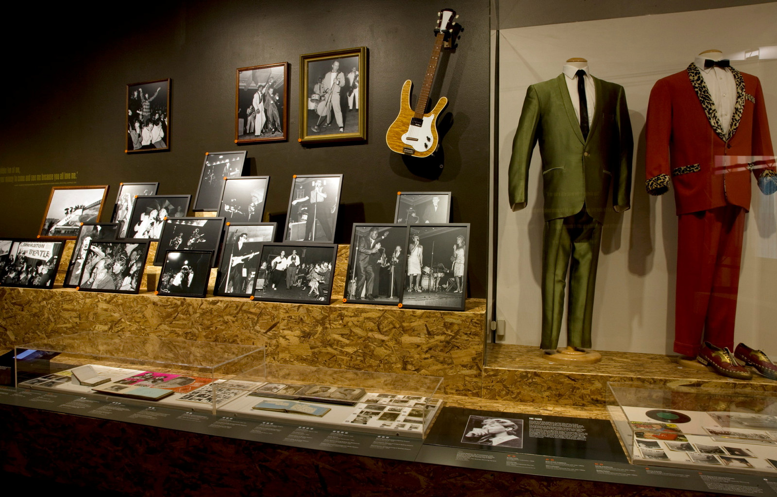 Interior view of exhibition space showing guitars, framed photographs and suits.