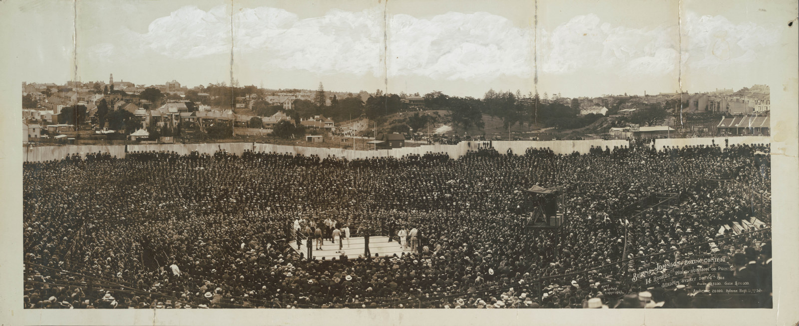 Wide angle image of a large crowd watching a boxing match. The crowd dresses almost all in black contrast with the white ring.