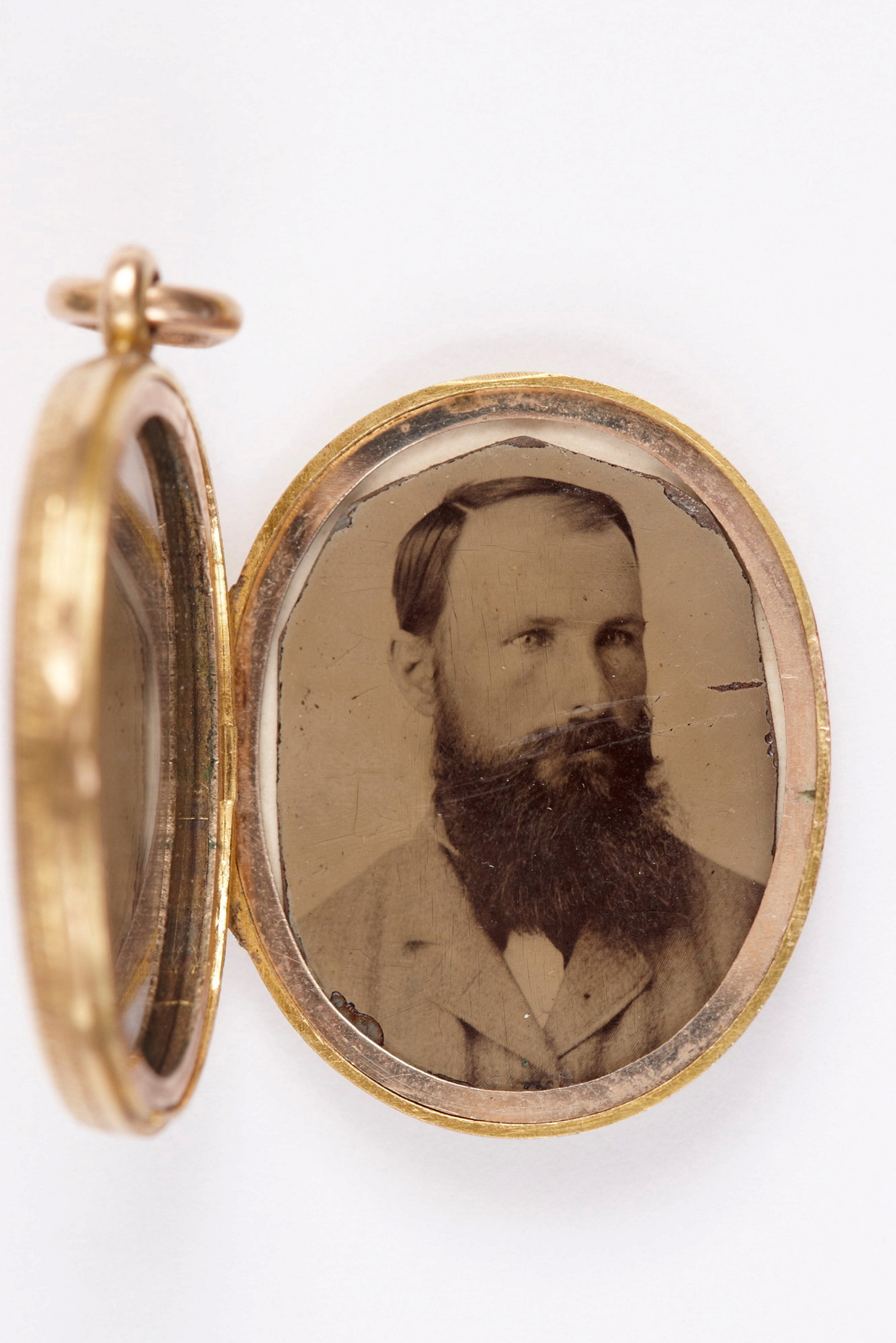 Handcoloured portrait tintype photograph of bearded man. Photograph sits within small oval gold locket, hinged at one side and attached with gold ring at top.