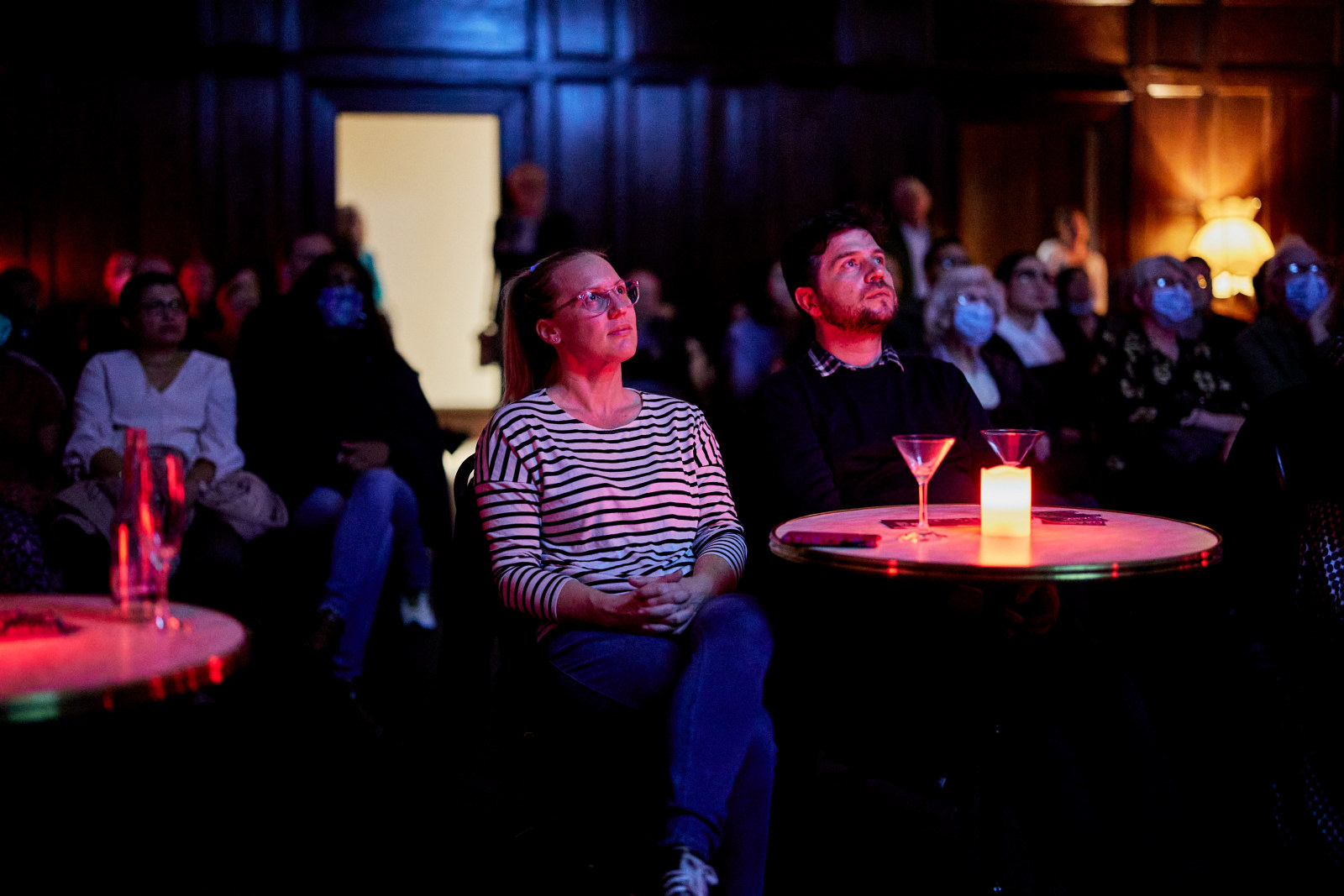Visitors look up, their faces illuminated by the light of the projected film. They are seated at round tables with candles and cocktails.