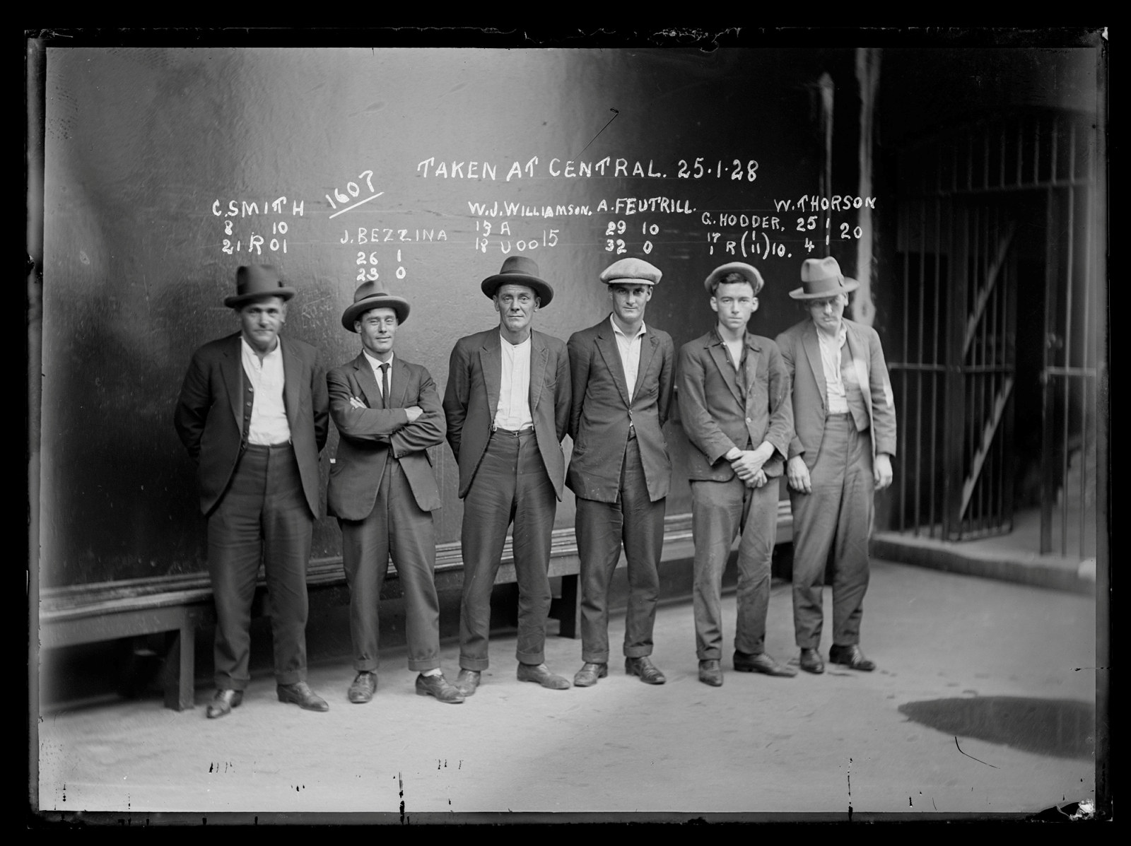 Black and white mugshot of six men standing in line, all with hats on.