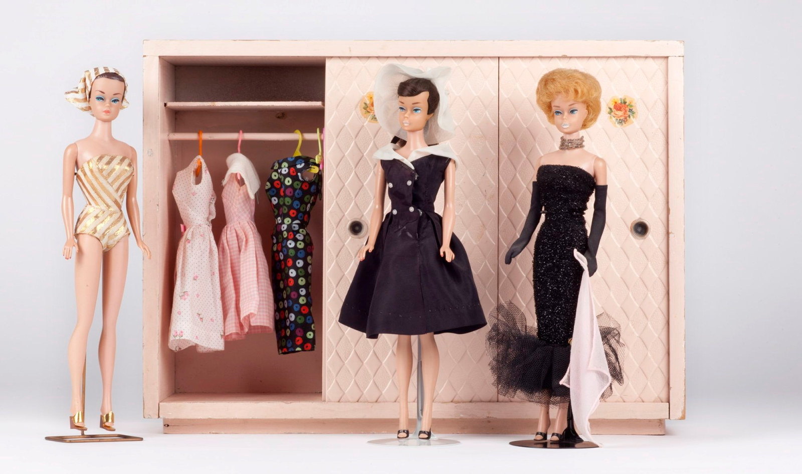 This is a colour photograph of three barbie dolls in various costumes standing in front of a 1950s style pink miniature wardrobe