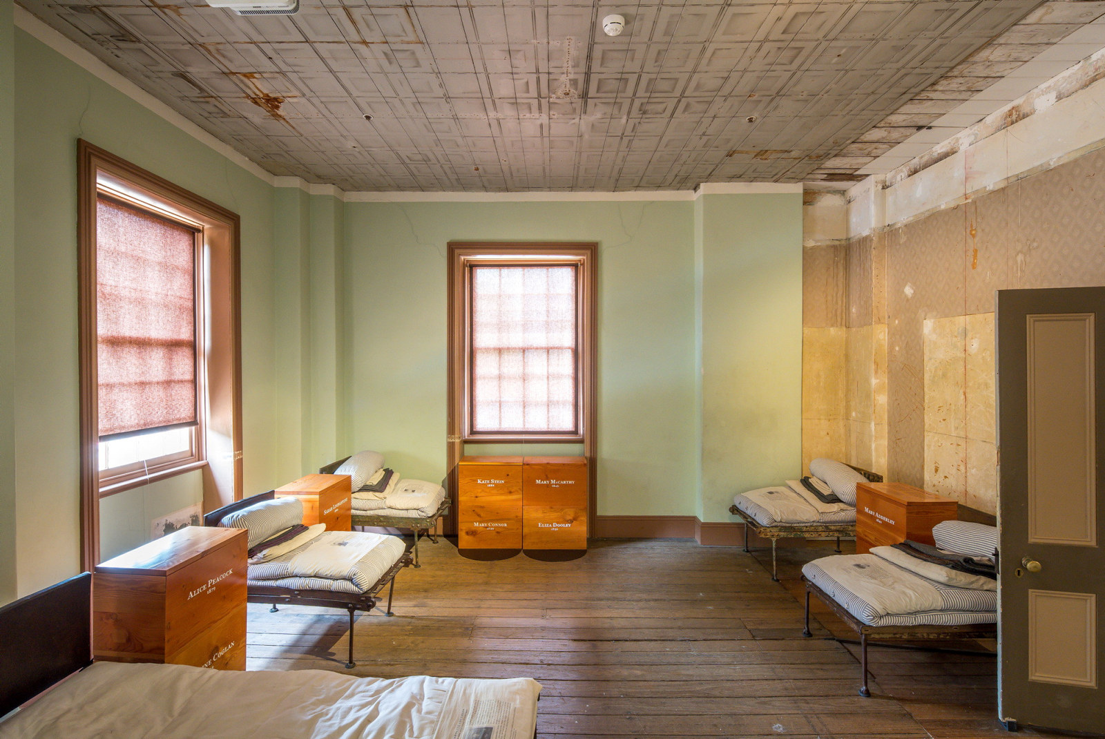Interior of wooden floorboarded room with natural light coming in window at end. Several beds placed around green painted walls.