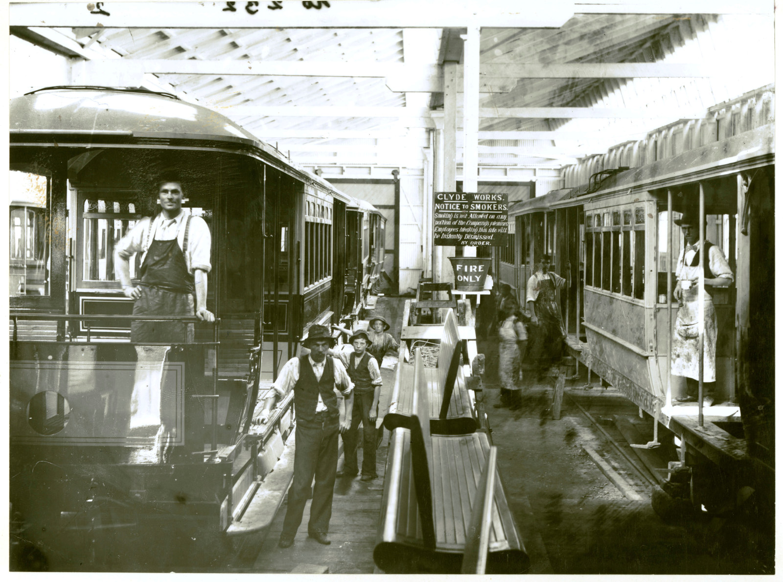 Trams being built in a workshop. Some of the numerous workers gather around looking at the camera.