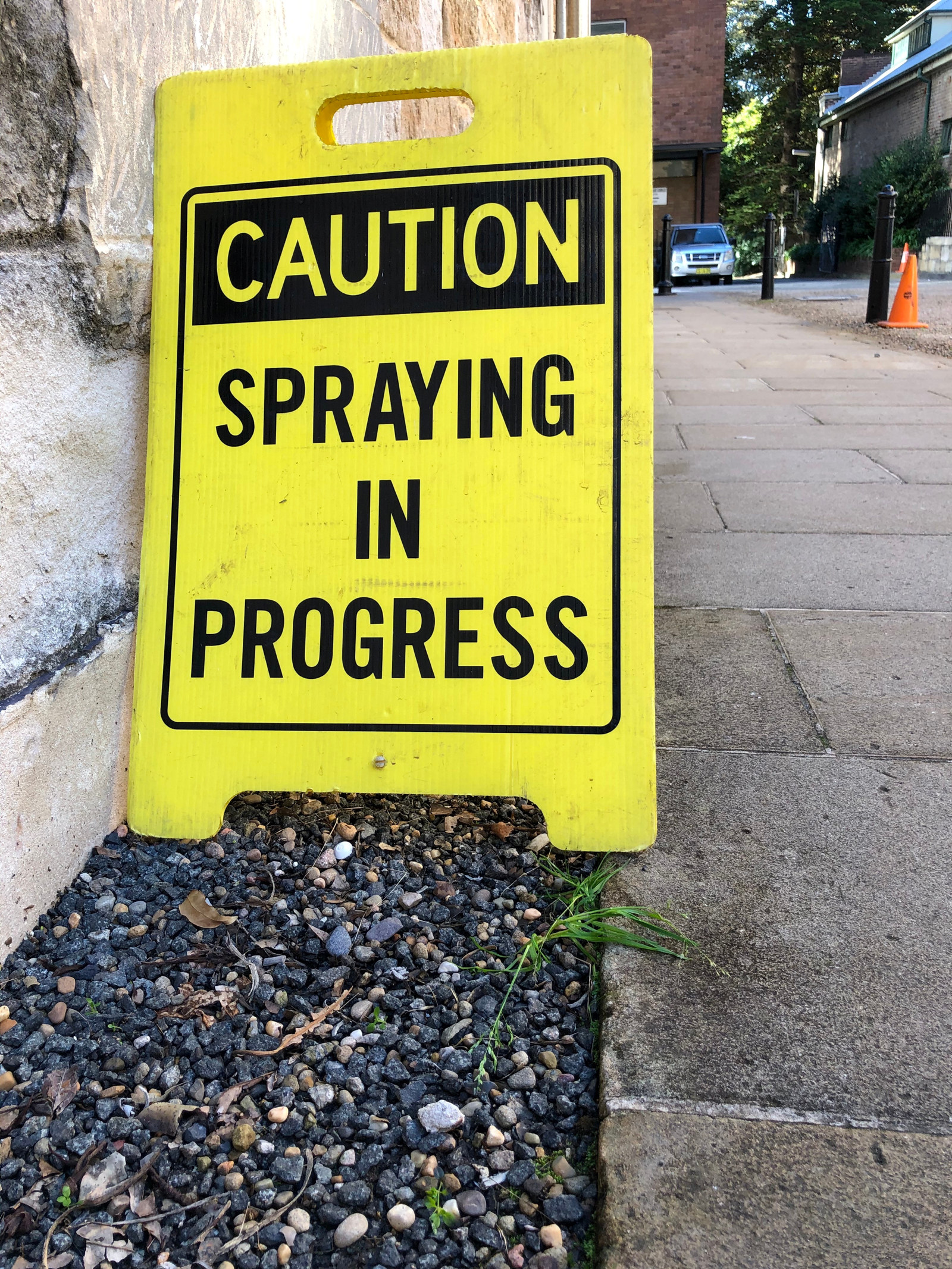 a yellow and black sign reads "caution spraying in progress" anlongside a sandstone wall and path