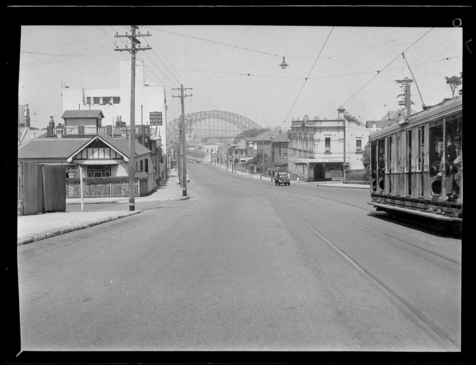 View along Pacific Highway toward Harbour Bridge with passing tram, general store, apartments.
