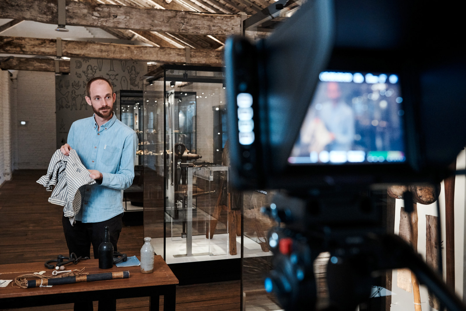 Carlin de Montfort, curator, holds up a convict shirt to the camera during a virtual excursion. He is standing behind a table of historical objects in the Meet the Convicts room on the top floor of the Hyde Park Barracks.