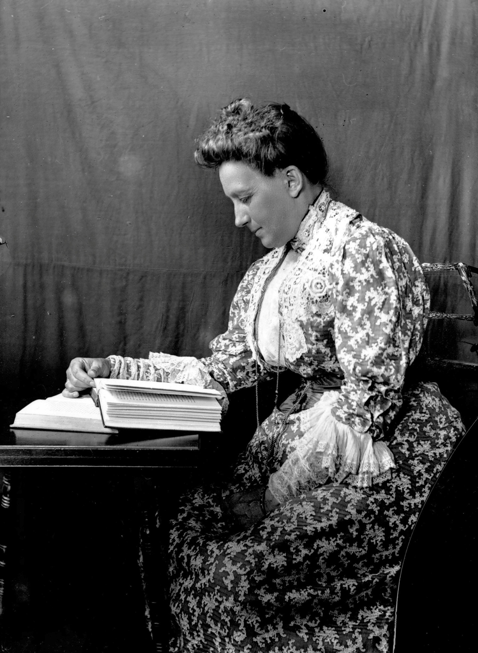 Photo of woman sitting at a desk reading, wearing a patterned high collar dress, facing the camera sideways