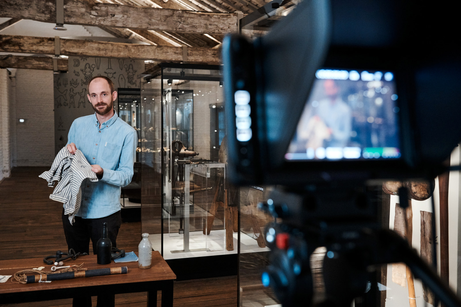 Carlin de Montfort, curator, holds up a convict shirt to the camera during a virtual excursion. He is standing behind a table of historical objects in the Meet the Convicts room on the top floor of the Hyde Park Barracks.