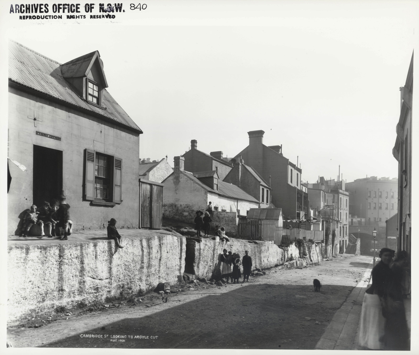 Photograph of street scene. Children and women stand outside or at the door of a row of small cottages.